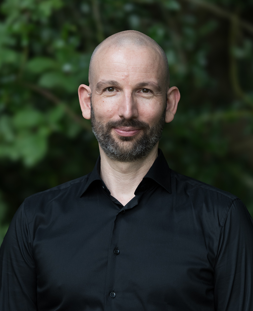 Potrait von Lars Hirschberger. Er trägt ein schwarzes Hemd, Bart und einer Glatze. Er schaut freundlich in die Kamera. Hintergrund sind verschwommene grüne Büsche zu sehen.
